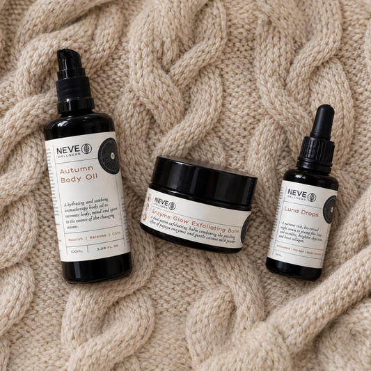 Three NEVE skincare products arranged on a chunky cream cable-knit blanket: a body oil bottle, an exfoliating scrub jar, and a dropper bottle.
