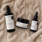 Three NEVE skincare products arranged on a chunky cream cable-knit blanket: a body oil bottle, an exfoliating scrub jar, and a dropper bottle.